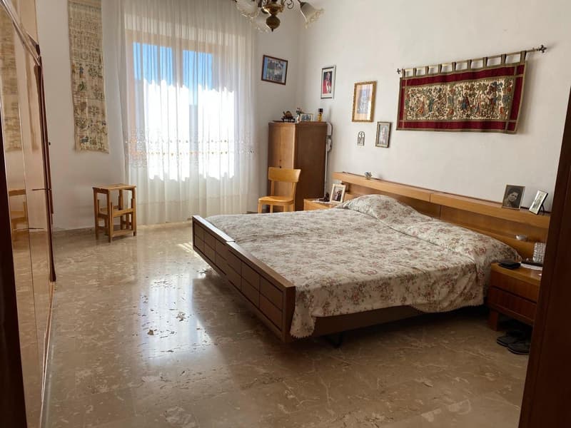 Spacious bedroom with hardwood floors, large windows, floral patterned bedding, wooden furniture including a dresser and nightstands, and decorative wall hangings.