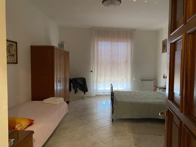 The image shows a spacious bedroom with a large bed, a wooden wardrobe, and a curtained window. The room has a tiled floor and appears to be well-lit.