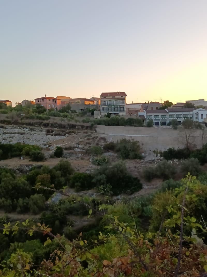 The image shows a residential area with several multi-story buildings and houses. The buildings have a mix of architectural styles, with some appearing more modern and others more traditional. The area is surrounded by greenery, including trees and vegeta
