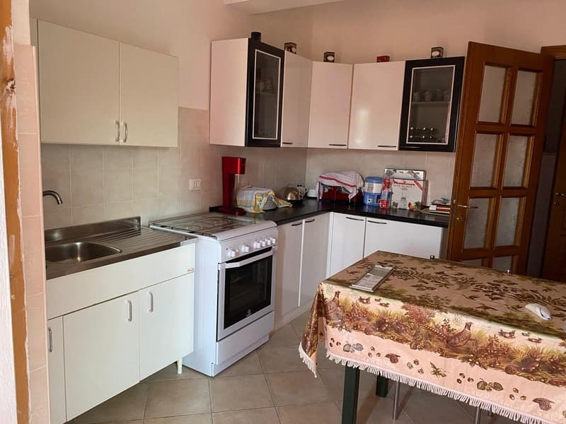 The image shows a kitchen with white cabinets, a stove, and a sink. There is a patterned tablecloth on a table in the kitchen. Various kitchen appliances and utensils are visible on the countertop.