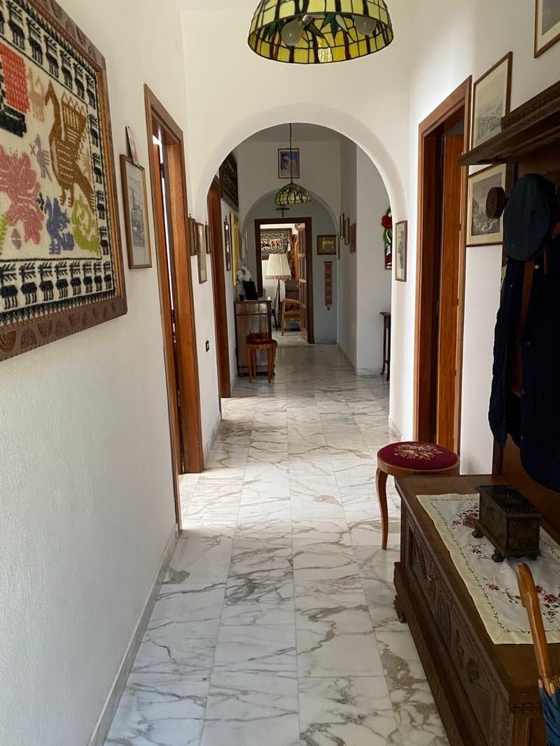 Marble tile flooring, arched doorways, decorative lighting fixture, framed artwork on the walls, wooden furniture pieces, and a red cushioned stool in the hallway
