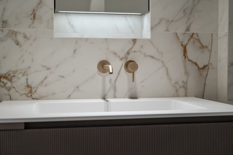 Bathroom with white marble walls, white sink, gold faucets, mirror with light