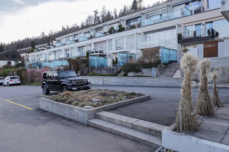 Modern building with multiple floors and balcony access, parking area with a few cars parked, landscaped area with steps leading to the entrance.
