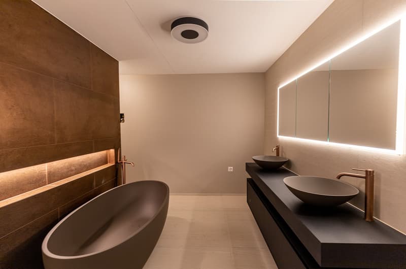 modern bathroom, beige tiled wall, bathtub, countertop, 2 sinks, faucets, mirror, LED light, electrical outlet