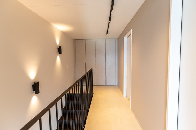 Corridor with white walls, yellow floors, black staircase, recessed lighting on ceiling, wall sconces