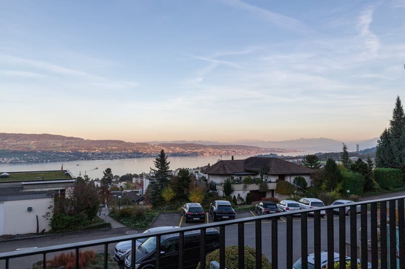 a property from a balcony with several cars parked and view of lake, mountains and buildings