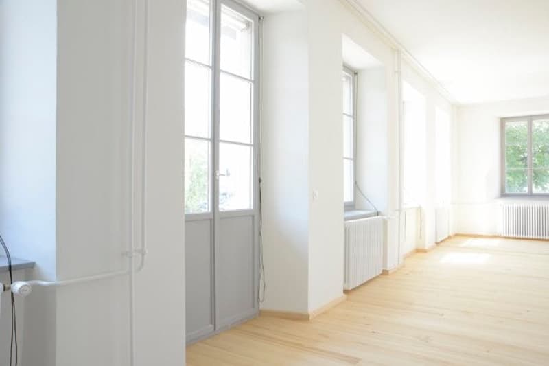 Empty room with white walls, wooden floor, large windows, and white radiators.