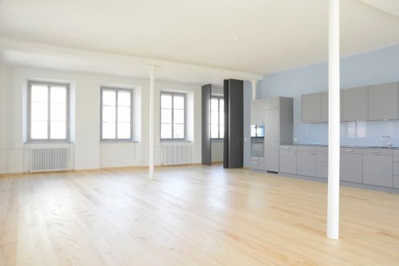 empty room, wooden floor, white walls, blue accent wall, large windows, kitchen area with cabinets and appliances