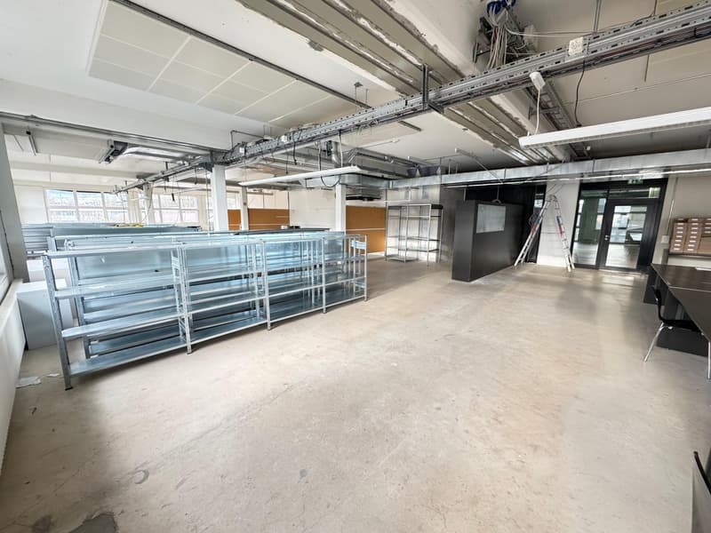Large open commercial space with concrete flooring, metal shelving units, and exposed ceiling with ventilation ducts and lighting. The space appears to be in a state of renovation or construction, with some equipment and materials visible.