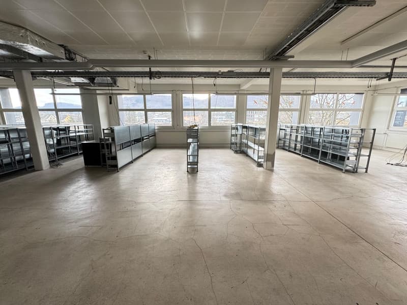 Large open space with concrete flooring, metal shelving units, and large windows providing natural light. The space appears to be a commercial or industrial setting, potentially a warehouse or storage facility.