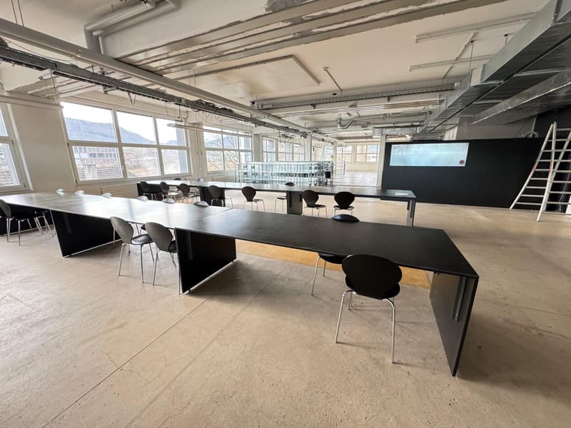 The image shows a large open office space with high ceilings, exposed beams, and large windows providing natural light. The space has a modern, industrial aesthetic with concrete floors and black furniture, including long tables and chairs. There is a pro