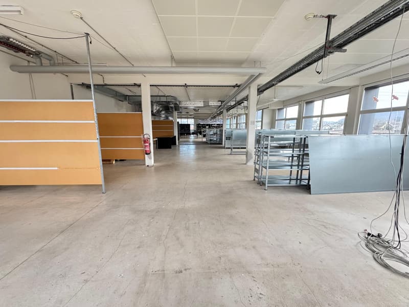 The image shows a large open office space with concrete floors, white ceiling tiles, and various equipment and furniture. The space appears to be under construction or renovation, with some partitions and shelving units visible. The windows provide natura