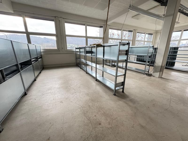 This image shows a large, open room with concrete flooring and several metal shelving units or racks lining the walls. The room has large windows that provide natural light, and there appears to be some equipment or machinery in the room as well.