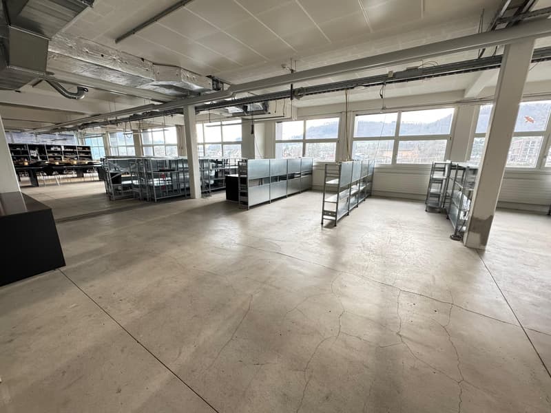 The image shows a large, open warehouse or storage space with high ceilings, concrete floors, and metal shelving units. The space appears to be empty, with no visible furniture or equipment. The windows provide natural light and offer a view of the surrou