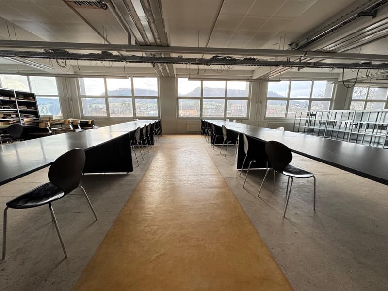 Large open office space with high ceilings, exposed beams, and large windows providing a panoramic view of the city. The space is furnished with rows of desks and chairs, suggesting it is used as a meeting or conference room.