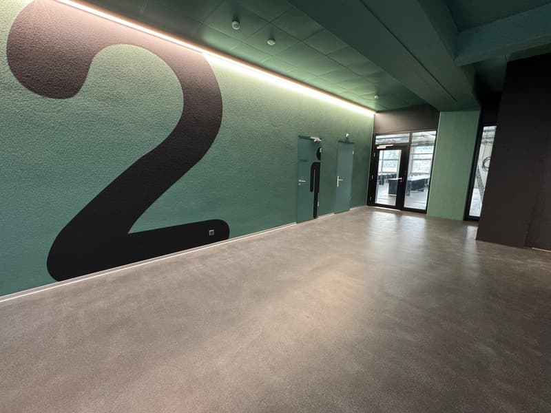 The image shows a large open office space with a green and black color scheme. The walls feature a large number '2' graphic design. The floor is covered in a light-colored concrete or tile material. The space has recessed lighting and glass doors leading