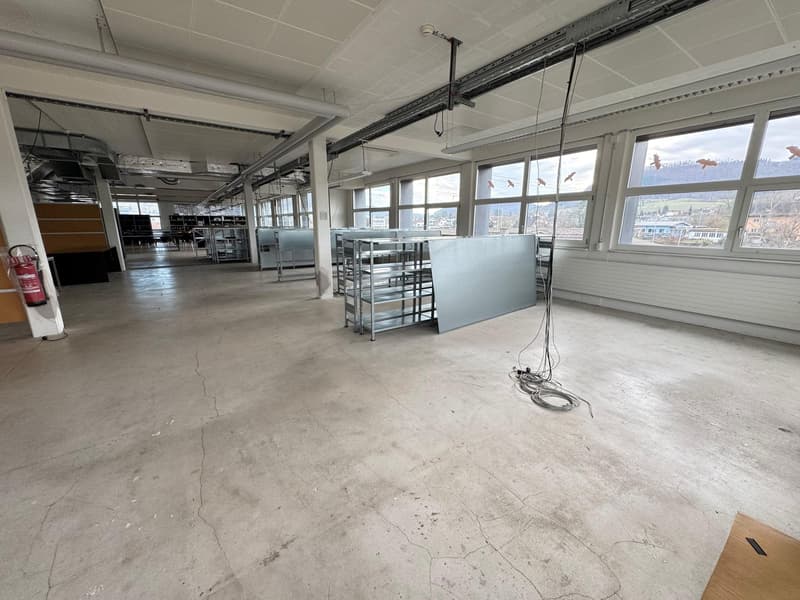 Large open commercial space with concrete flooring, metal shelving units, and overhead lighting. The space has large windows providing natural light and a view of the surrounding area.