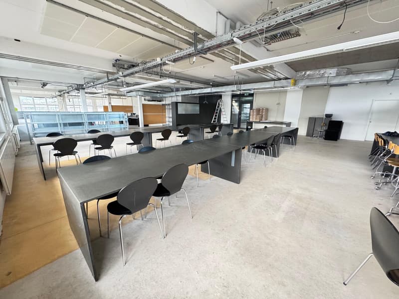 Large open-plan office space with concrete floors, exposed ceiling beams, and black chairs and tables. The space appears to be set up for meetings or collaborative work, with multiple workstations and a central conference table.