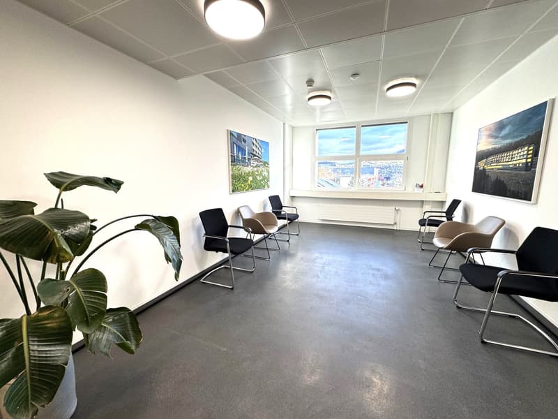 This image shows a modern office space with a minimalist design. The room has a concrete floor, white walls, and a suspended ceiling with recessed lighting. There are several black and beige chairs arranged in a semi-circle, suggesting a meeting or waitin