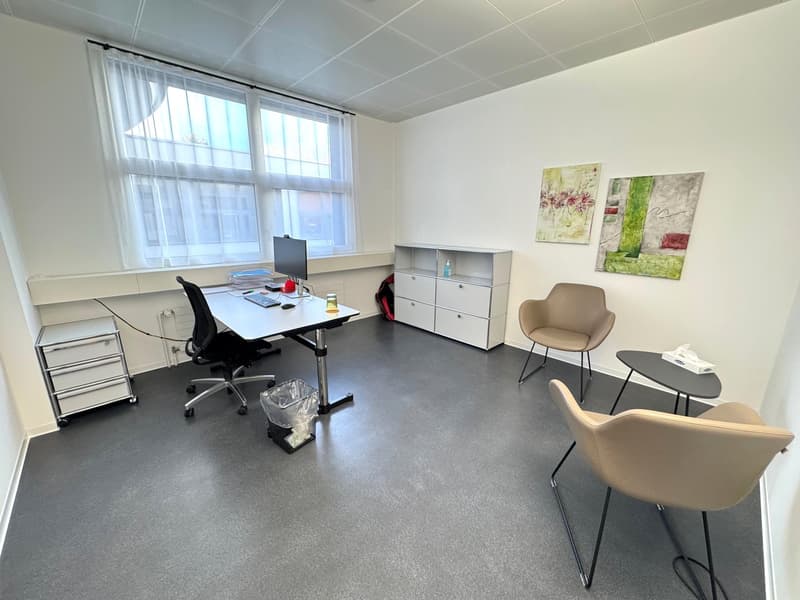 The image shows a modern office space with a desk, computer, office chair, storage cabinets, and artwork on the walls. The room has large windows that provide natural light, and the flooring is a dark gray color.