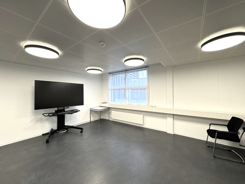 The image shows a modern office space with a large TV mounted on the wall, a desk with a computer monitor, and two chairs. The room has a minimalist design with white walls, a gray floor, and recessed lighting fixtures in the ceiling.