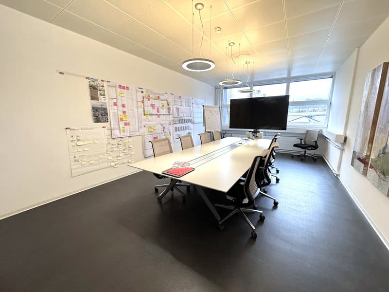 Empty meeting room, white walls, gray flooring, long desk, large monitor, swivel chairs, bulletin boards, natural light from large window