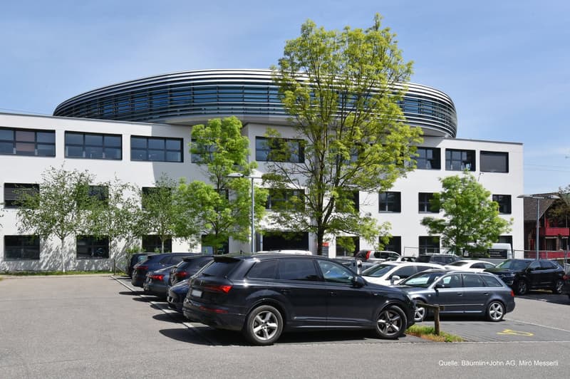 modern office building with glass facade, parking lot with multiple cars, trees
