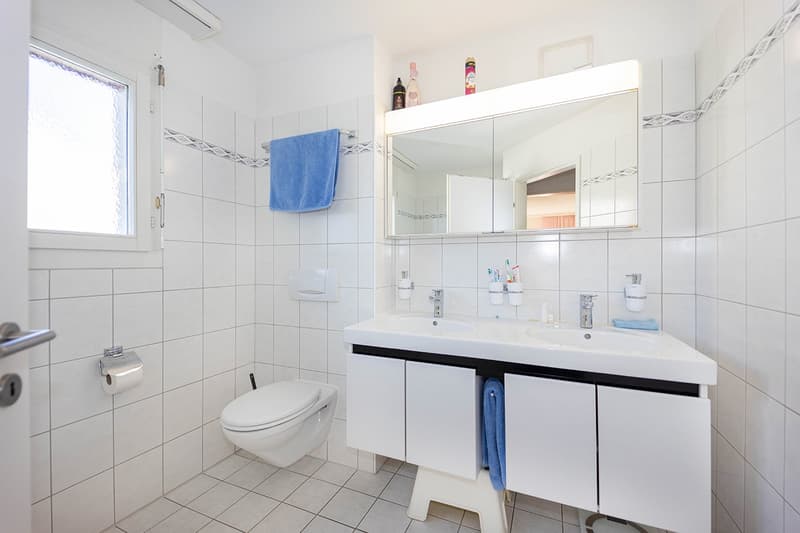 white tile, double sink with faucets, cabinet, large mirror, small window, white toilet, towel rack
