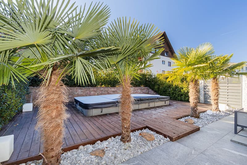 A wooden deck with a jacuzzi in the center, surrounded by white pebbles and large palm trees.