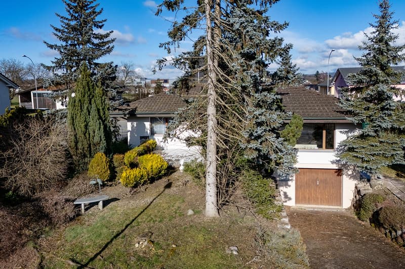 Detached house, gable roof, white walls, brown roof, wooden garage, trees, bushes, bench