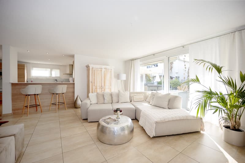 large living area, white couch, round silver table, plant, bar stools, cabinets, glass windows