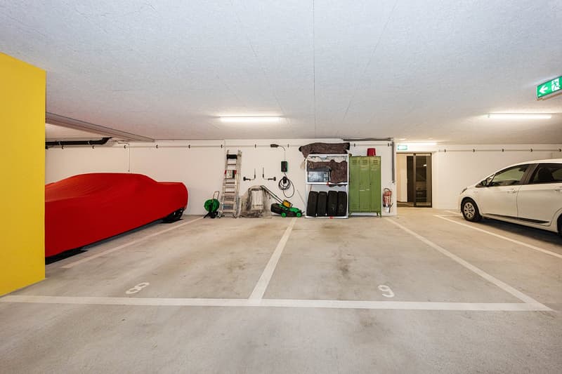 underground parking area, two parking spots, one car covered with a red cover, one car parked, green lockers, fire extinguisher, ladder, and other tools