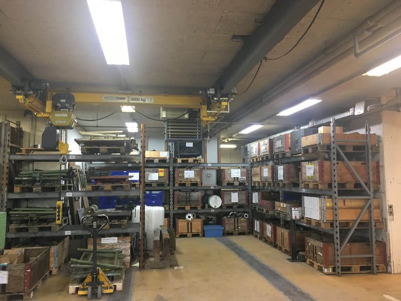 A large storage room filled with various items on wooden and metal pallets, suspended ceiling cranes, ceiling-mounted lighting, and shelves filled with materials.