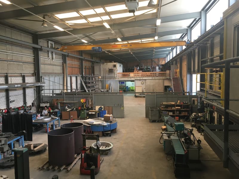 Warehouse with a variety of tools and machines, including heavy machinery, conveyor belts, and a yellow crane. A man in a safety vest and helmet is present, working with tools and equipment.
