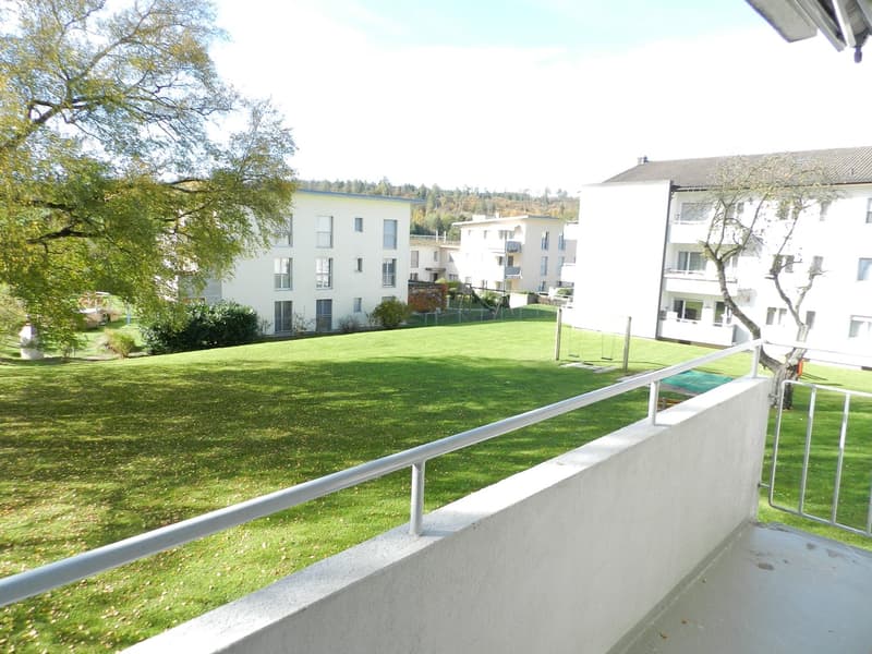 Exterior view, balcony with metal railing, grass lawn, playground with swings, buildings, and trees