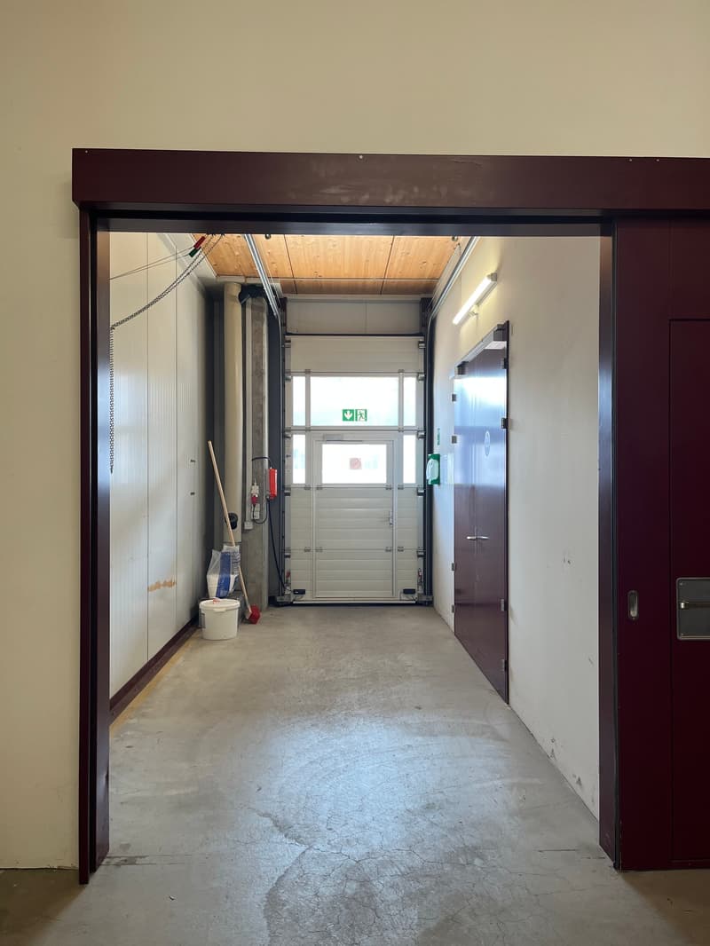 The image shows a hallway or entryway area of a building. The hallway has concrete flooring and appears to be a commercial or industrial space. There is a large garage door visible at the end of the hallway, indicating this may be a garage or storage area