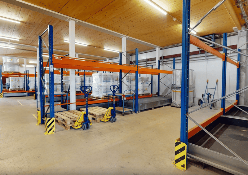 Warehouse with large shelving units, trolleys, pallets, and illuminated by ceiling lights.