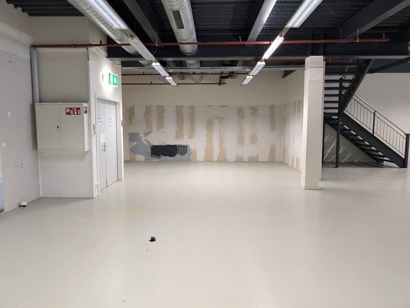 An empty room with white walls, white floor, exposed pipe and wires on ceiling, door, and stair
