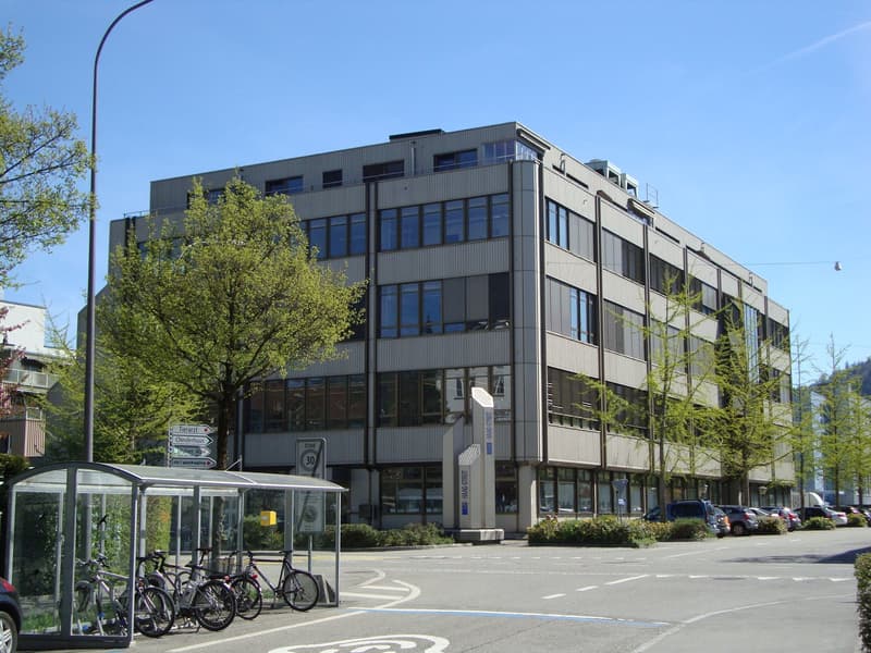 large building with multiple stories and many windows, large bushes and trees in front, bicycles parked at the side, cars parked in front