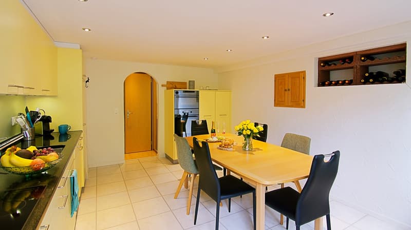 Spacious open-plan kitchen and dining area with yellow walls, wooden cabinets, stainless steel appliances including a dishwasher, a dining table with chairs, and a wine rack on the wall