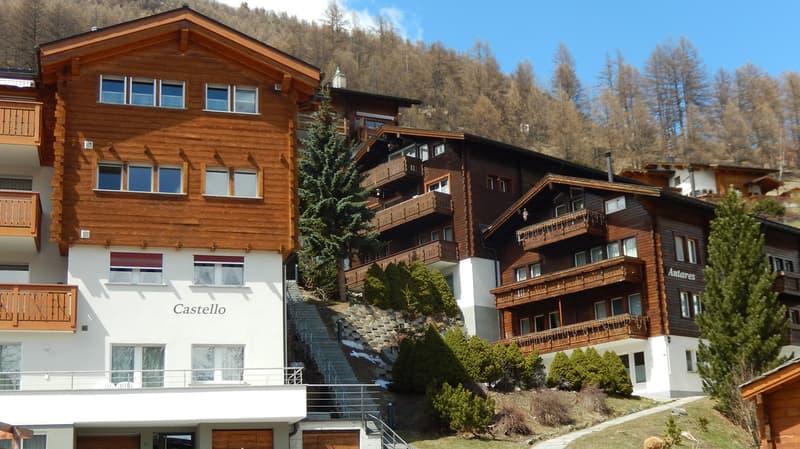 Modern alpine architecture, wooden exterior, multiple terraces, large windows, set against a mountainous backdrop.