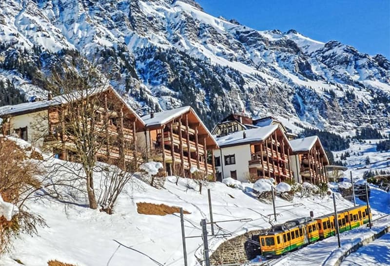 Wooden chalets, snow-covered mountains, yellow train on tracks, winter scenery