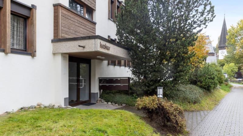 White building with wooden trims, glass door, mailbox, grassy area, trees, pathway
