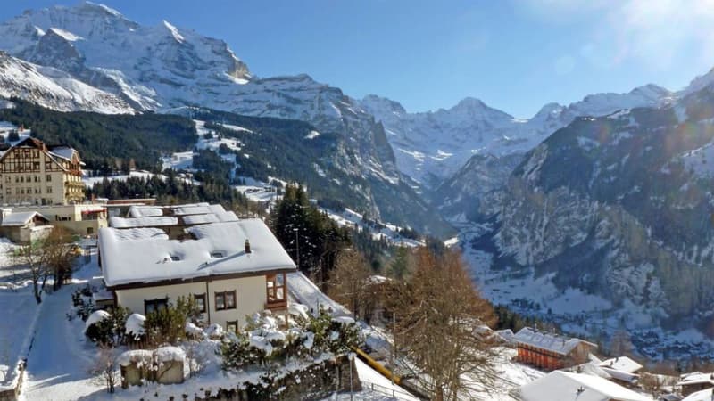 House with snow-covered roof, surrounded by snow-covered mountains, trees, and other houses