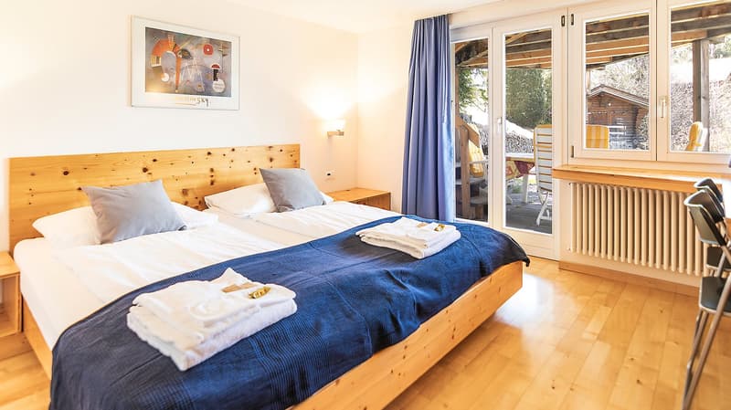 double bed, wooden headboard, white linen, blue blanket, white towels, nightstands, picture frame, sliding glass doors, blue curtains, wooden floor, radiator