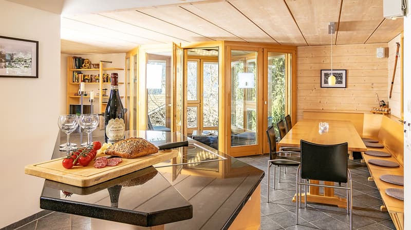 kitchen with dining area, wooden floor, large windows, wooden table, wooden chairs, wooden shelves, wine bottle, wine glasses, bread, tomatoes, prosciutto, view of the outside