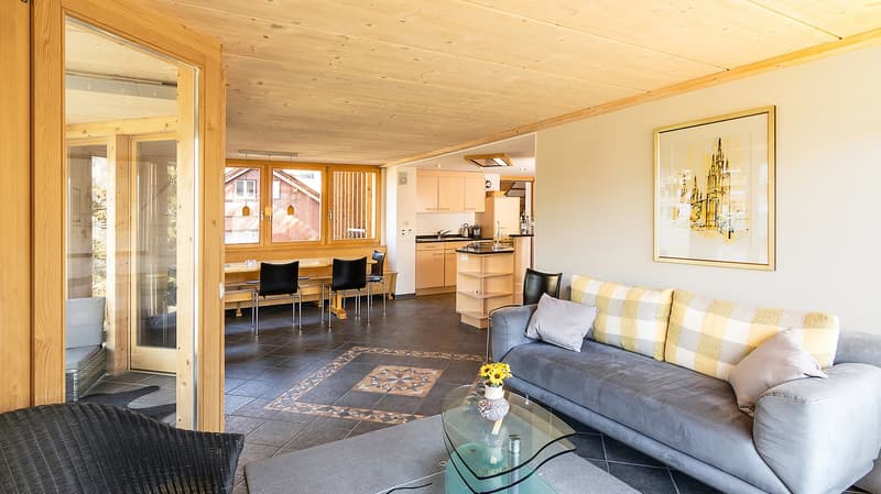 living room with wooden ceiling, gray couch, glass table, yellow flowers, dining table, kitchen, windows