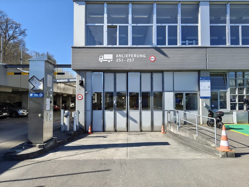 A building with a sign that reads 'Anlieferung 251-257', a parking area with a barrier, and a scooter parked in front.