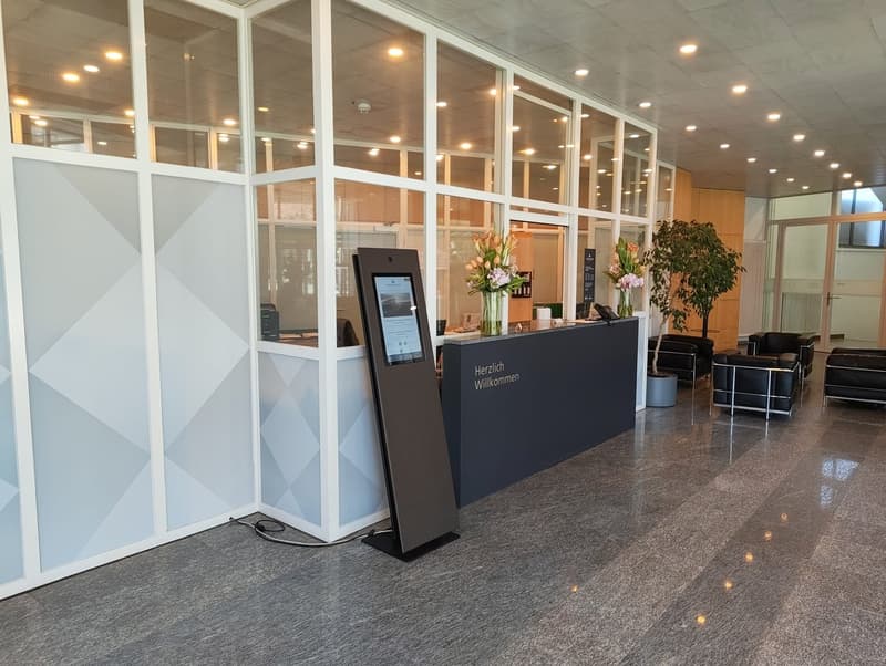 Reception desk with monitor, flowers, black chairs, gray floor, white walls, glass partitions