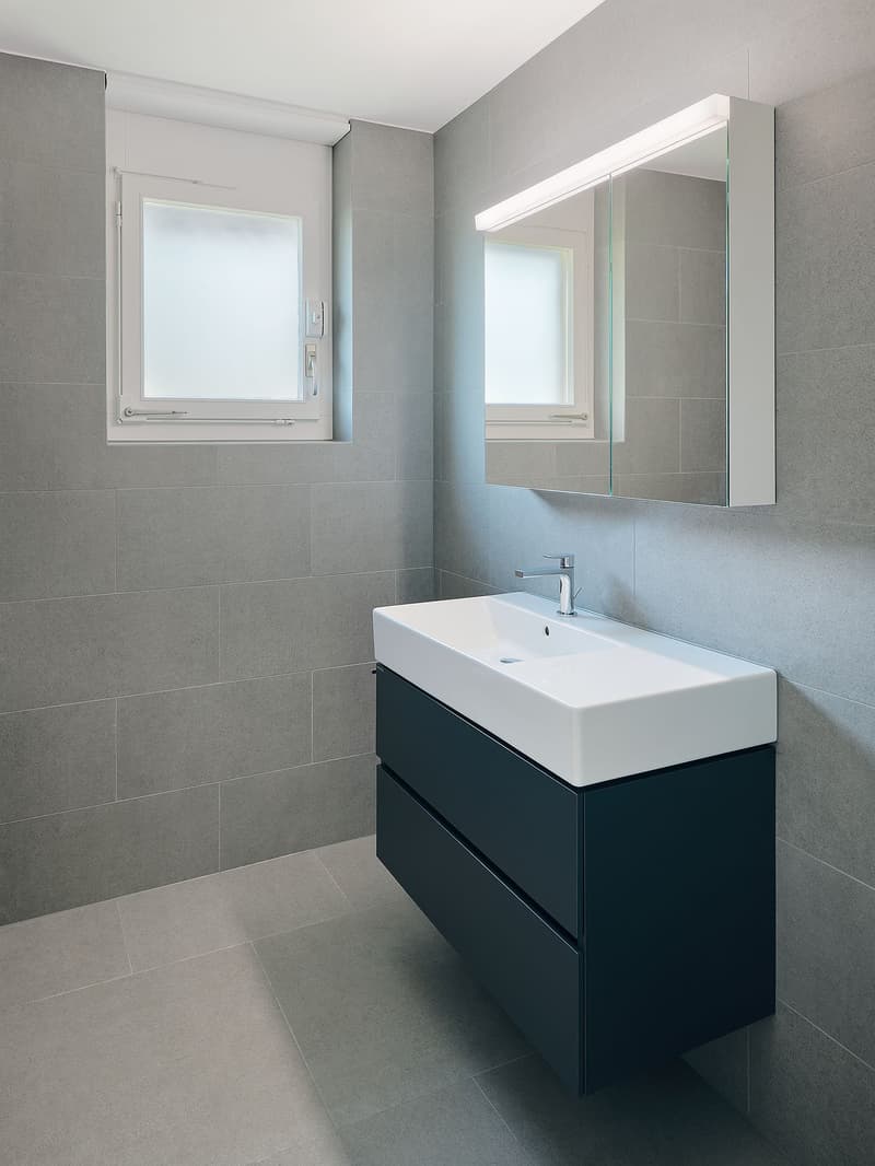 Bathroom with grey tiles, white sink, black cabinet, mirror with light, and white window.